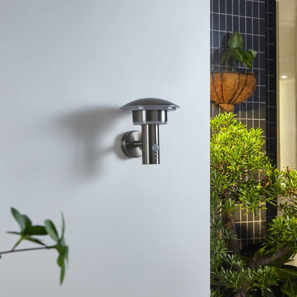 Moderne Außenwandleuchte aus Edelstahl mit Bewegungsmelder, montiert an einer weißen Wand neben grünen Pflanzen.