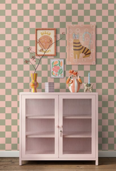 Vlies-Tapete mit Schachbrettmuster vor einem rosa Metallschrank mit Glastüren, dekoriert mit Vasen und Bildern an der Wand.
