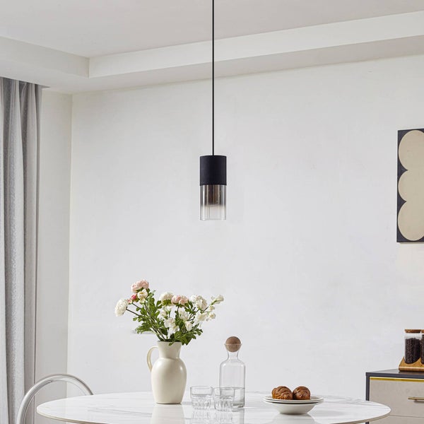 Moderne, zylindrische Pendelleuchte mit schwarzem Gehäuse und Glasschirm über einem Esstisch in einem hellen, modern eingerichteten Raum.