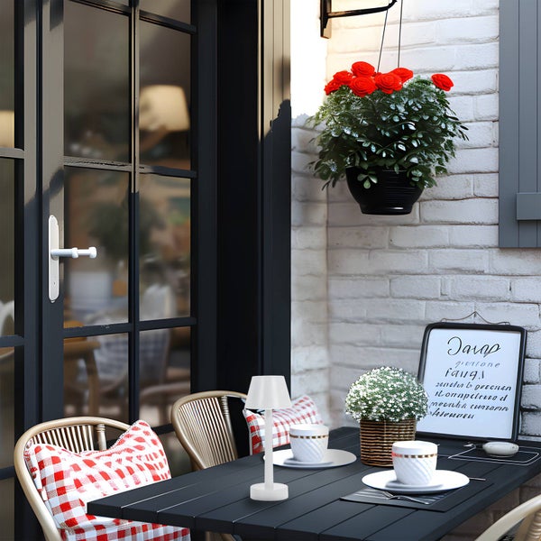 Weiße LED-Tischleuchte auf einem dunklen Gartentisch mit Stühlen und roten Kissen vor einer hellen Ziegelwand.