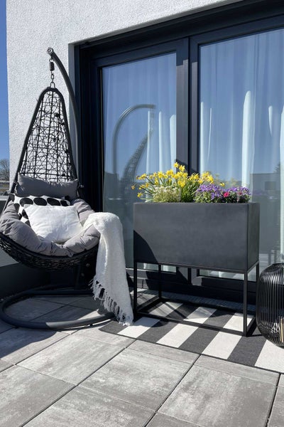 Helle, moderne Terrasse mit schwarzem Hängesessel aus Polyrattan, rechteckigem Pflanzkasten mit Blumen und gestreiftem Teppich auf grauen Fliesen.