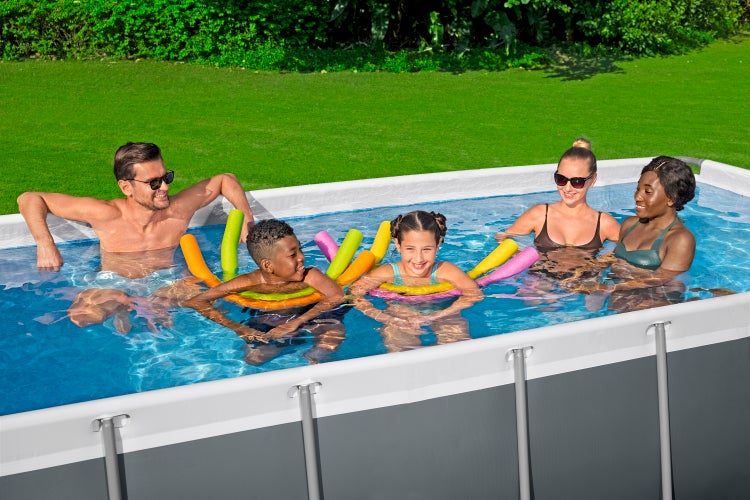 Zwei Erwachsene und zwei Kinder in einem rechteckigen Aufstellpool mit Metallrahmen und Poolnudeln im hellen Garten auf einer Rasenfläche.