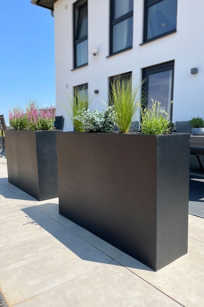 Zwei rechteckige Pflanzkübel als Raumteiler auf einer hellen, organisierten Terrasse, bepflanzt mit Gräsern und Blumen.