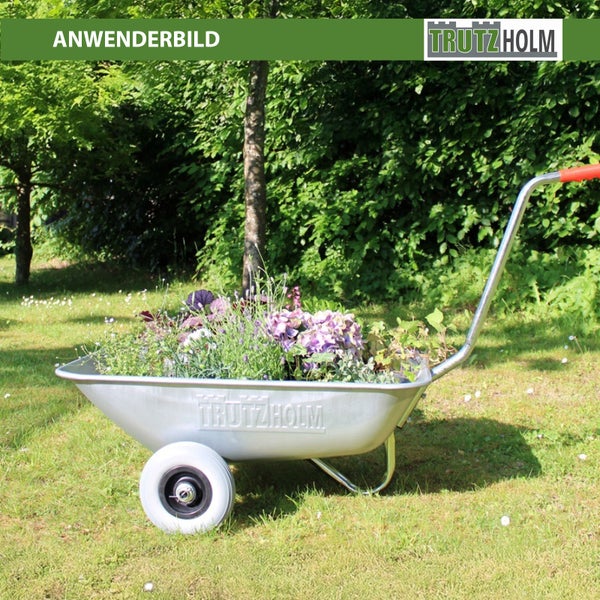 Eine metallene Trutzholm Gartenkarre gefüllt mit Blumen auf einer Rasenfläche in einem Garten.