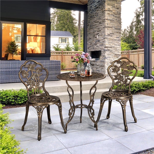 Dreiteiliges Bistro-Set aus Aluminiumguss in antik-bronze auf einer hellen, geordneten Terrasse. Tisch und zwei Stühle mit dekorativem Rosenmuster.
