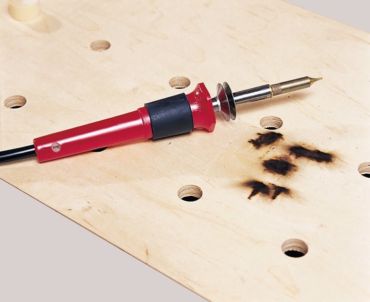 Ein elektrischer Brandmalkolben für Holzbearbeitung liegt auf einer Holzplatte mit Brandmarkierung.