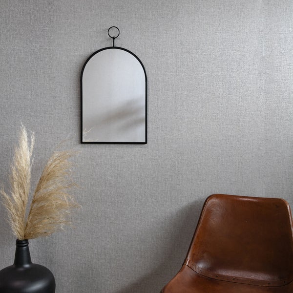 Wandspiegel in Bogenform mit schwarzem Metallrahmen und Aufhängering an einer strukturierten Wand neben einem Sessel und einer Vase mit Ziergras.