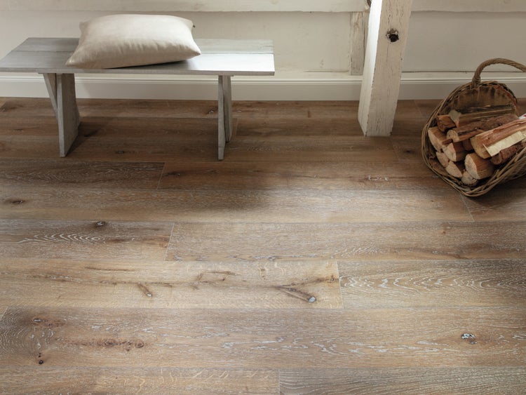Heller Raum mit rustikalen Holzbodendielen, einer grauen Holzbank mit Kissen und einem Weidenkorb mit Brennholz.