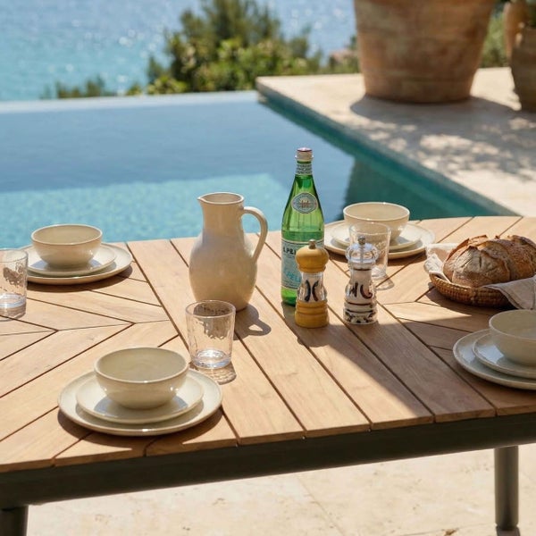 Gedeckter Gartentisch aus Holz mit Fischgrätenmuster neben einem Pool, mit Keramikgeschirr, Gläsern und einem Krug.