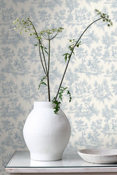 Vliestapete mit blauem Toile-de-Jouy-Muster, das ländliche Szenen zeigt, dekoriert mit einer weißen Keramikvase und einer Schale auf einem Tisch.
