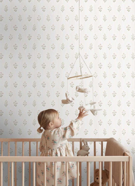 Hell gestaltetes Kinderzimmer mit gestreifter Vliestapete im Blumenmuster, ein Kind greift aus einem Holzgitterbett nach einem Mobile.