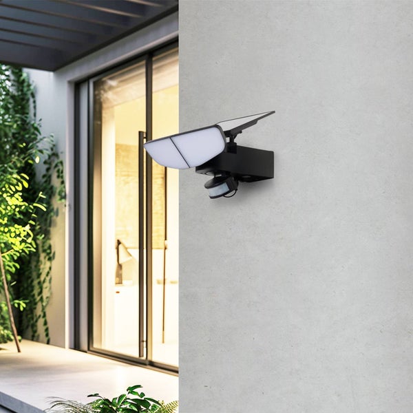 Schwarzer LED-Solaraußenstrahler mit Bewegungsmelder und Solarpanel an einer hellgrauen Wand neben einer gläsernen Terrassentür montiert.