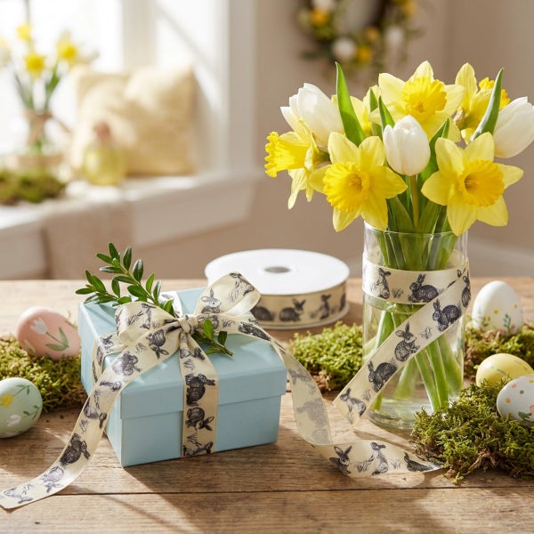 Osterdekoration mit Narzissen in Glasvase und Geschenkbox verziert mit Osterhasen Geschenkband auf Holztisch.