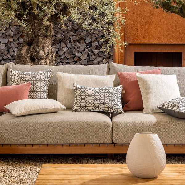 Modernes Outdoor-Lounge-Sofa mit Holzgestell und grauen Polstern, dekoriert mit Musterkissen auf einer hellen Terrasse mit Olivenbaum und Brennholz.