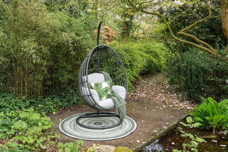 Hängesessel in Eiform mit Metallgestell, Kissen und Decke auf einem runden Teppich im Garten, umgeben von Bambus und Pflanzen.