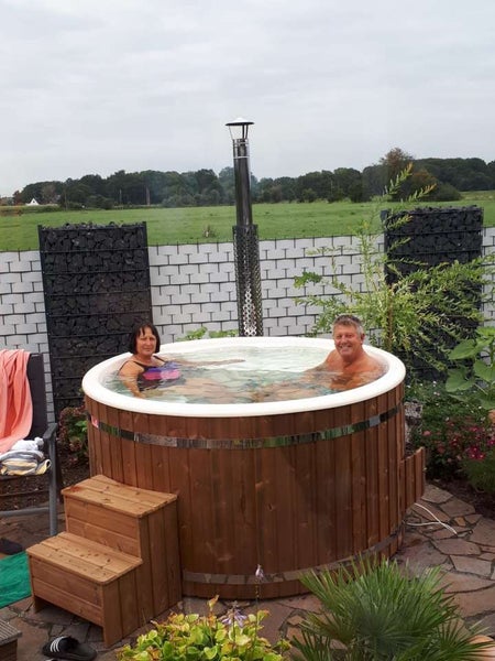 Zwei Personen entspannen in einem hölzernen Badezuber mit Ofen im Garten.
