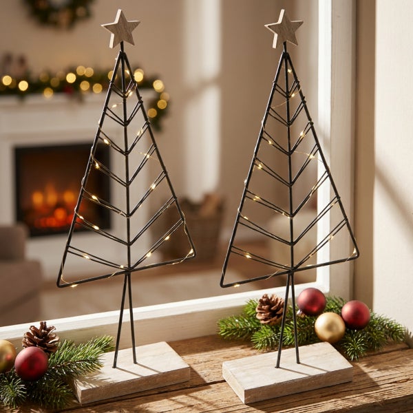 Zwei dekorative Weihnachtsbaume aus Metall mit LED Beleuchtung und Holzsockel als weihnachtliche Fensterdekoration auf einem Holztisch.