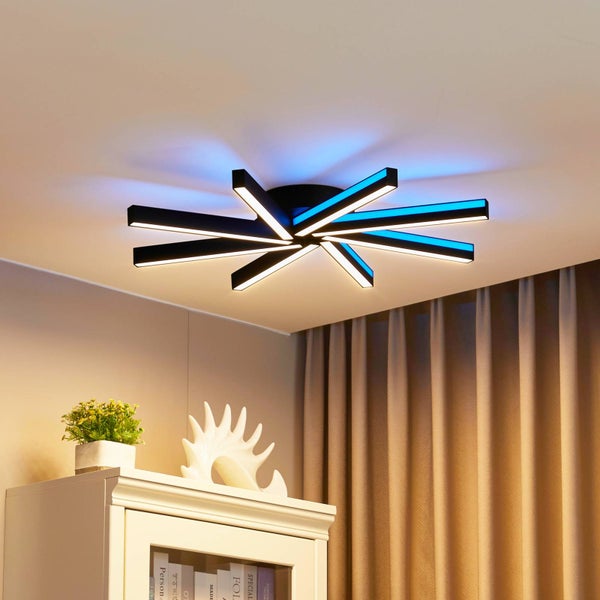 Moderne LED-Deckenleuchte mit sternförmig angeordneten schwarzen Stäben und farbigem Lichteffekt an der Decke in einem hellen Wohnraum.