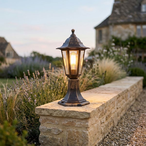 Klassische Sockelleuchte für den Außenbereich in Antik-Optik, montiert auf einer Steinmauer im Garten mit leuchtendem Leuchtmittel.