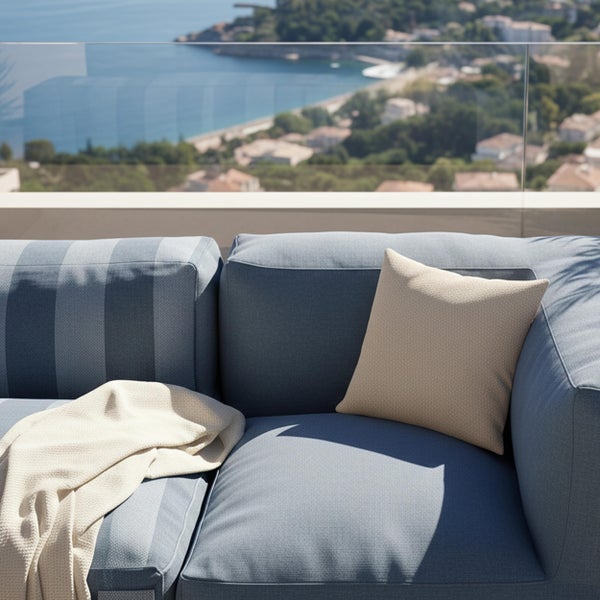 Modulares Outdoor Sofa in Blau mit gestreiftem Sitzkissen, beigem Dekokissen und beiger Decke auf einem Balkon mit Meerblick.