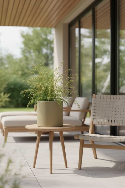 Moderner Außenbereich mit rundem Holzbeistelltisch, olivgrünem Pflanztopf mit Ziergras und einem geflochtenen Holzstuhl auf einer hellen Terrasse.