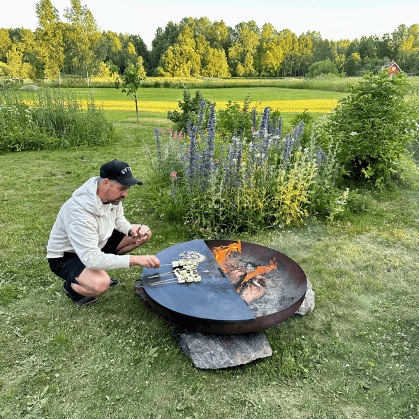 Eine Person grillt Spieße auf einer Feuerschale mit Grillplatte in einem weitläufigen Garten mit Wiesen und Sträuchern.