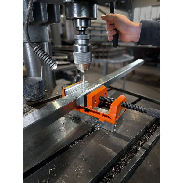 Maschinenschraubstock in Orange fixiert ein Metallprofil beim Bohren mit einer Standbohrmaschine.