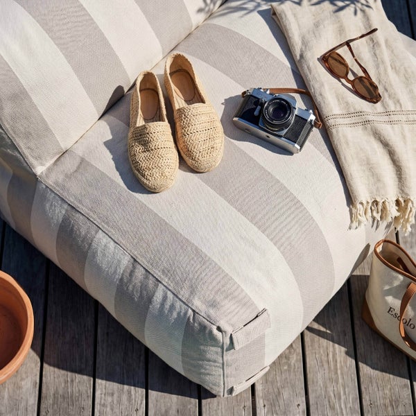Gestreifter Outdoor-Sitzsack mit Espadrilles, Kamera und Decke auf einer Holzterrasse im Sonnenlicht.