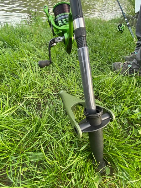 Angelrutenhalter im Boden verankert, hält eine Angelrute mit Rolle auf einer Wiese am Wasser.