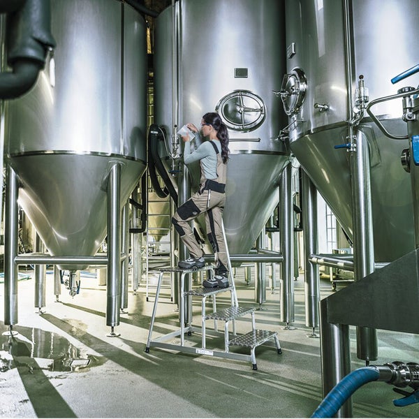 Eine Person steht auf einer Aluminium-Stufenleiter vor großen Edelstahltanks in einer Industrieanlage und führt eine Prüfung durch.