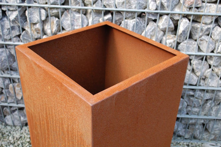 Quadratischer Pflanzkübel aus Metall mit Rostoberfläche vor einer Gabionenwand.