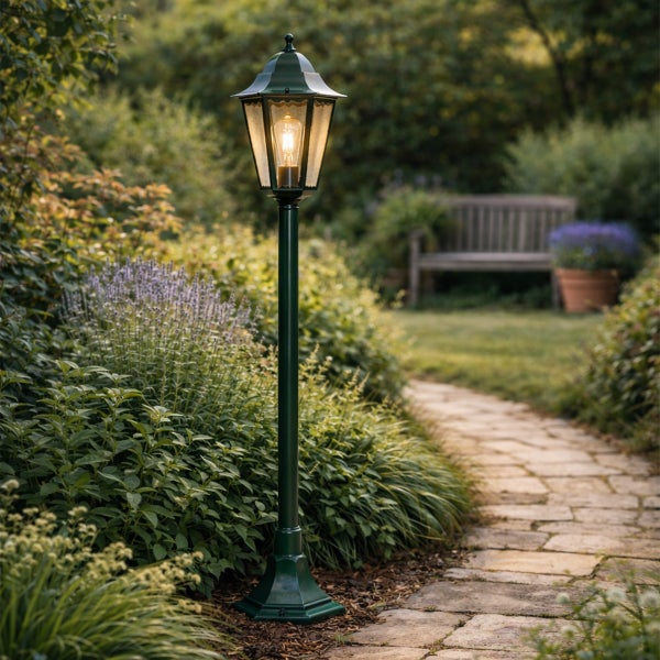 Klassische Gartenlaterne auf grünem Pfosten neben einem Steinpfad in einem hellen, bepflanzten Garten mit einer Holzbank im Hintergrund.