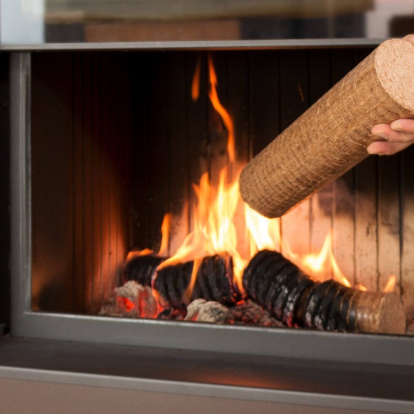Eine Hand legt einen zylindrischen Holzbrikett in einen brennenden Kamin