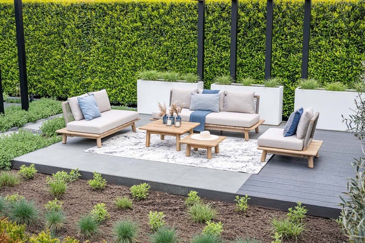 Gartenlounge aus Holz mit beigen Polstern, blauen Akzenten, Sofas, Sessel und zwei Tischen auf einer Terrasse mit Teppich vor einer Hecke.