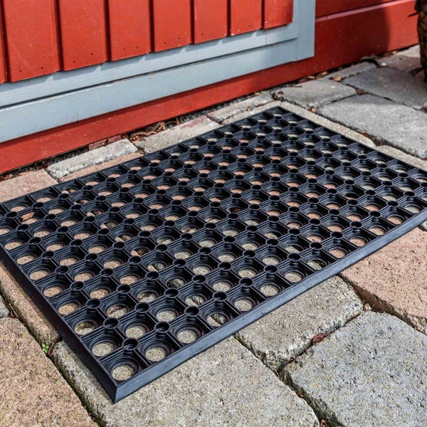 Schwarze Fußmatte mit Lochmuster vor einer roten Holzwand