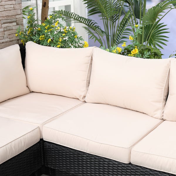 Nahaufnahme eines dunklen Rattan-Ecksofas mit cremefarbenen Kissen, umgeben von grünen Pflanzen und gelben Blumen im Garten.