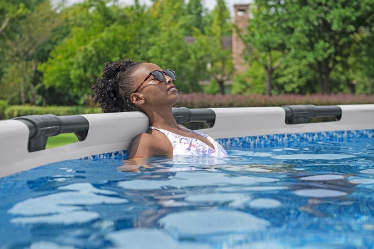 Eine Frau mit Sonnenbrille entspannt in einem hellen Gartenpool und lehnt ihren Kopf an den breiten Beckenrand.