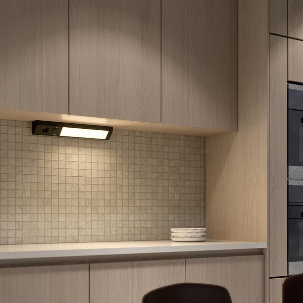 Schwarze Unterbauleuchte mit Schalter unter einem Küchenschrank aus Holz. Das Licht beleuchtet eine Arbeitsplatte und einen Fliesenspiegel.