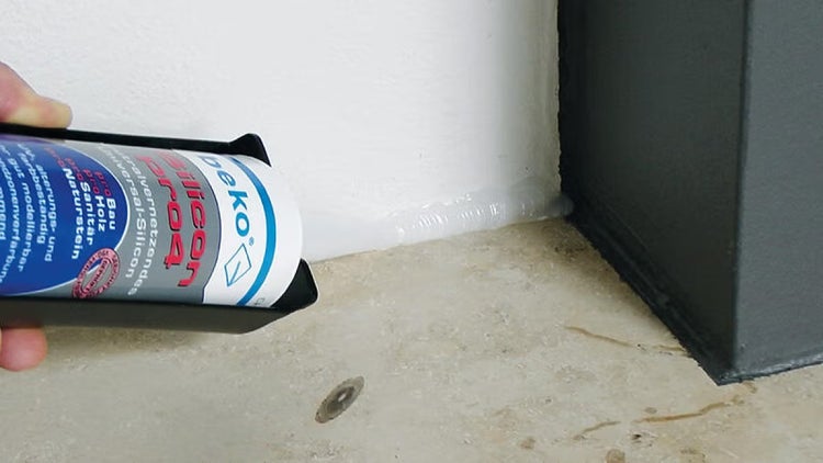 Eine Hand trägt mit einer Kartuschenpresse Beko Silicon Pro4 Dichtstoff als Fugenabdichtung entlang einer Wand und eines Natursteinbodens auf.