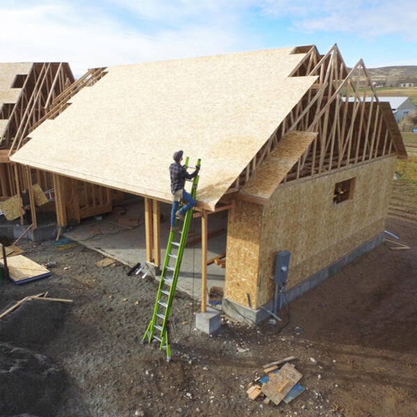 Bauarbeiter auf einer grünen Leiter montiert Holzbauplatten auf dem Dachstuhl eines Rohbaus aus Holz.