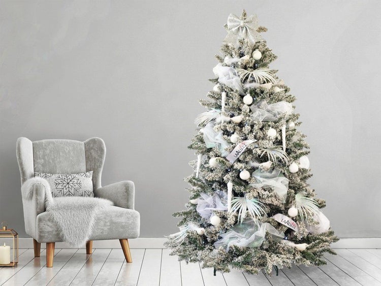 Künstlicher, beschneiter Weihnachtsbaum geschmückt mit Kugeln und Schleifen neben einem Sessel in einem winterlich dekorierten Raum.