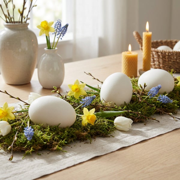 Osterdekoration mit Eiern, Narzissen, Traubenhyazinthen und Moos auf einem Holztisch, dekoriert mit zwei brennenden Bienenwachskerzen.