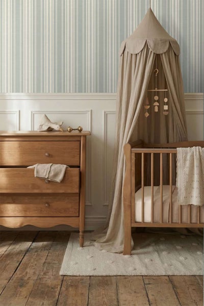 Ein Kinderzimmer mit gestreifter Tapete, hölzerner Kommode, Kinderbett mit Betthimmel und Spieluhr, gestaltet in einem klassischen, hellen Stil.