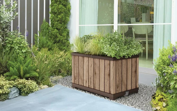 Rechteckiger Pflanzkasten in Holzoptik mit dunklem Rand, bepflanzt mit Grünpflanzen, auf einer Kiesterrasse vor einer Glastür.