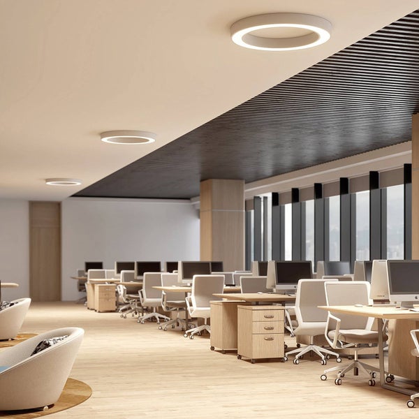 Moderne Bürolandschaft mit ringförmigen Deckenleuchten über Schreibtischen mit Monitoren und Bürostühlen in einem hellen, organisierten Raum.