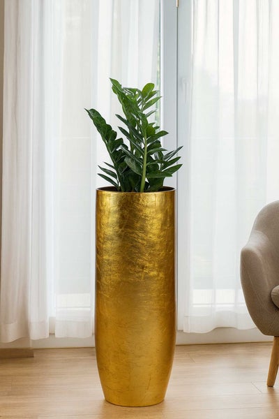 Hoher zylindrischer Pflanzkübel mit goldener Struktur und grüner Pflanze in einem hellen Raum mit Holzfußboden und Vorhängen.