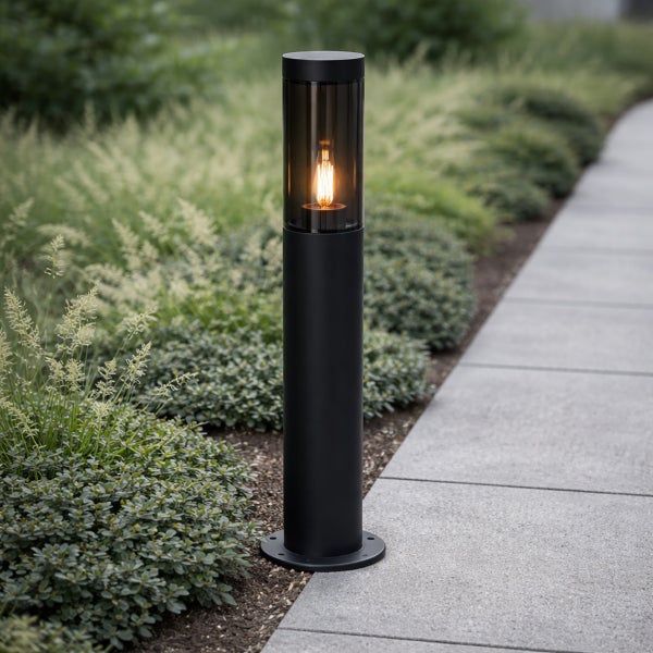 Schwarze zylindrische Wegeleuchte aus Metall mit Glaszylinder und brennendem Filament-Leuchtmittel neben einem gepflasterten Gartenweg.