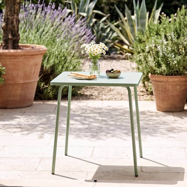Kleiner hellgrüner quadratischer Gartentisch aus Metall auf einer hellen Steinterrasse, dekoriert mit Blumen und Snacks, umgeben von Pflanztöpfen.