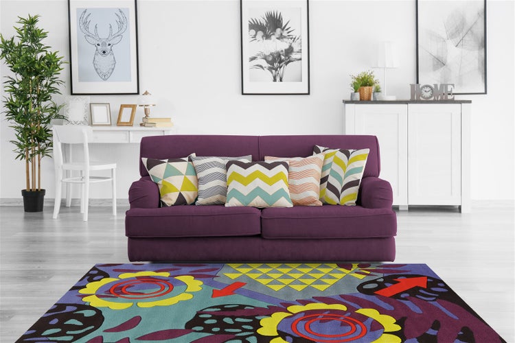 Wohnzimmer mit lilafarbenem Sofa, dekoriert mit gemusterten Kissen, vor einem bunten Teppich mit abstraktem Blumenmuster und geometrischen Formen.