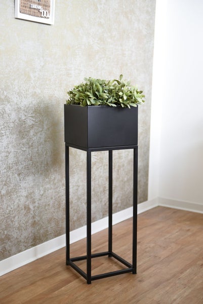 Moderner hoher Blumenständer aus Metall mit rechteckigem Pflanzkasten auf Holzboden vor einer gemusterten Wand.